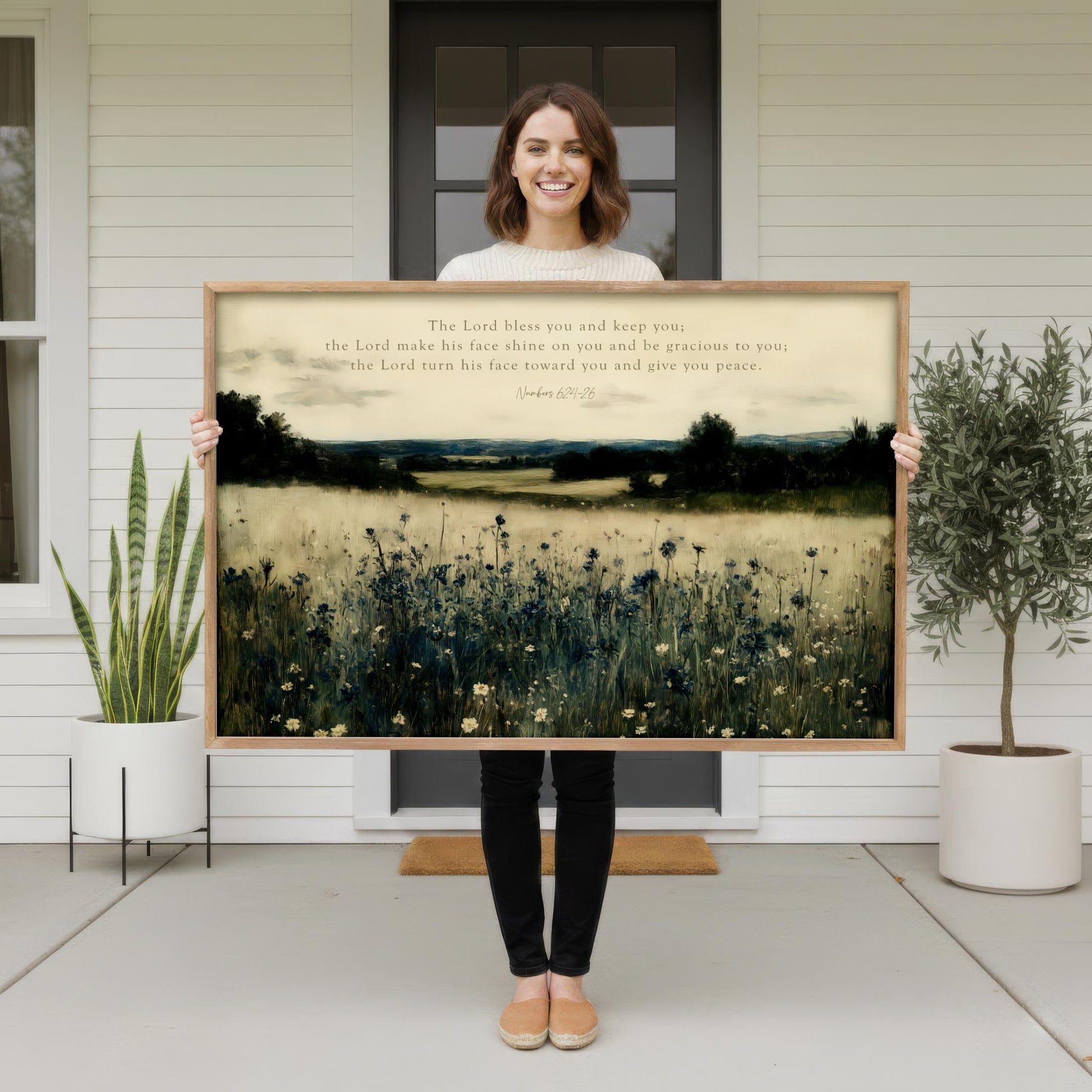 A woman stands in front of a white house, holding a large framed painting of a field with flowers.