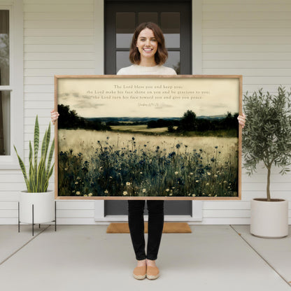 A woman stands in front of a white house, holding a large framed painting of a field with flowers.