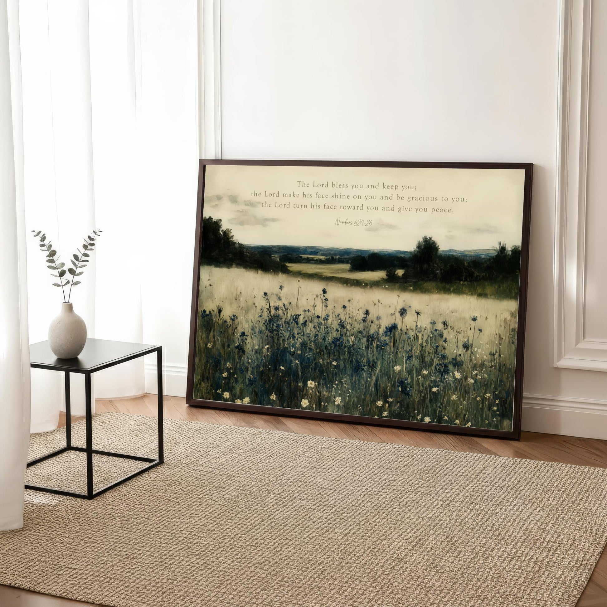 a framed painting of a field with tall grass and wildflowers, placed on a wooden floor in a room with a rug and a small table with a vase of greenery.