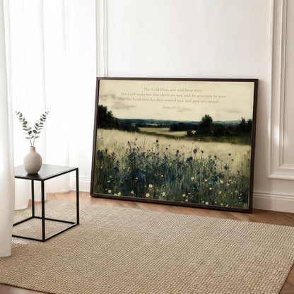 a framed painting of a field with tall grass and wildflowers, placed on a wooden floor in a room with a rug and a small table with a vase of greenery.