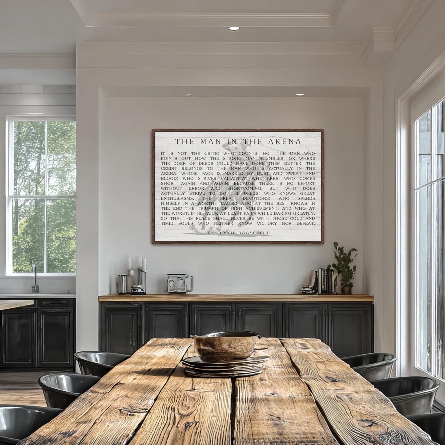 a rustic wooden dining table with a bowl on top, surrounded by black chairs. Above the table, there is a framed quote in black text that reads "The man in the arena".