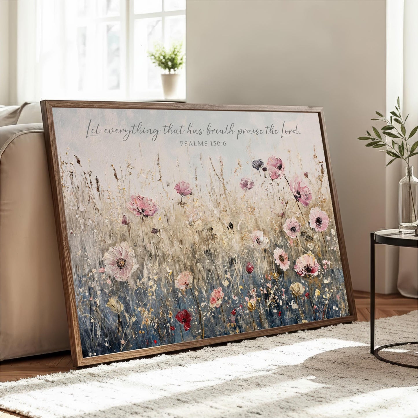 A framed painting of a field of flowers is displayed on a wooden stand in a living room, with a potted plant and a vase nearby.