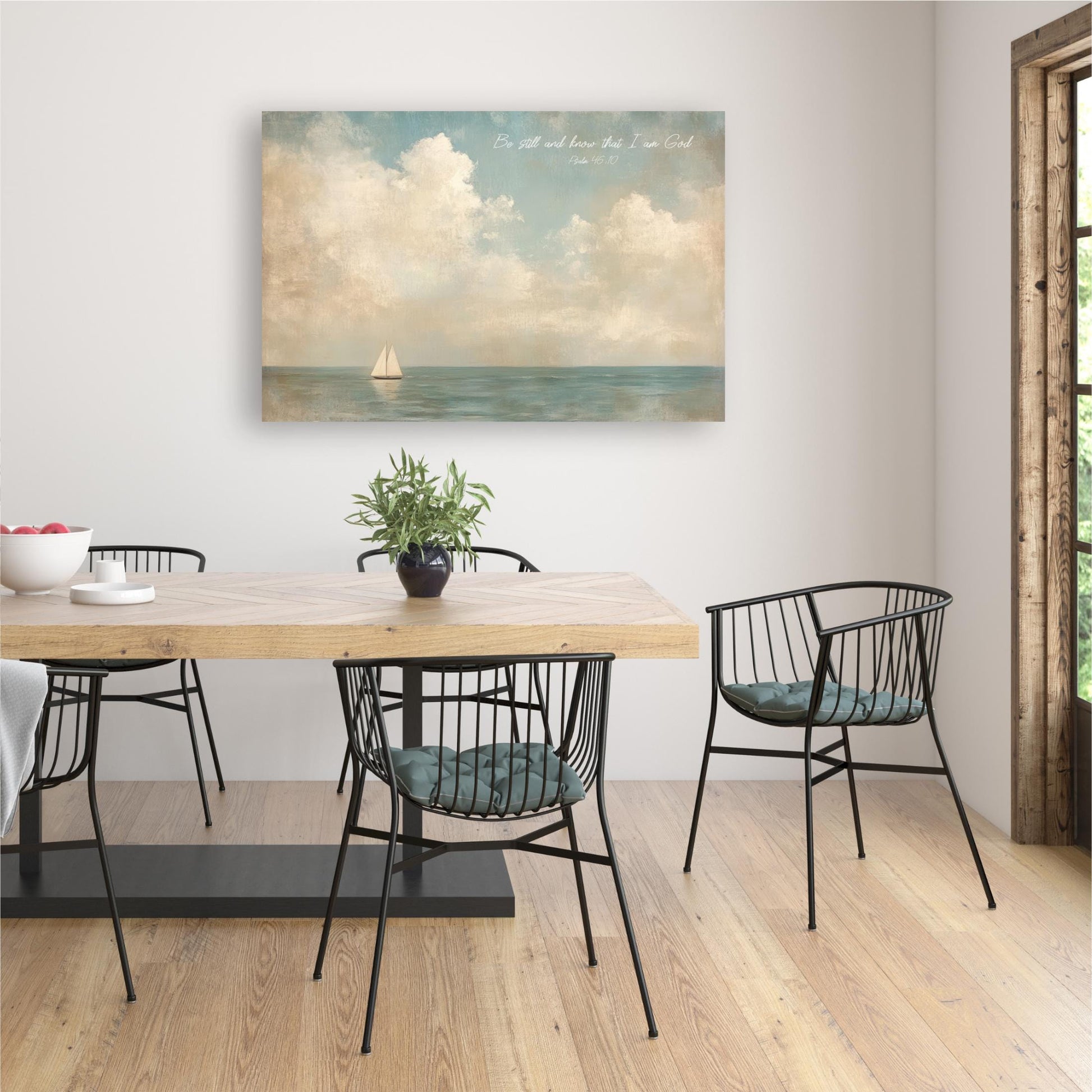 a dining room with a wooden table and black metal chairs. Above the table, there is a painting of a sailboat on the ocean. The painting is framed and hangs on the wall. The table is set with a bowl and a plant, creating a cozy and inviting atmosphere.