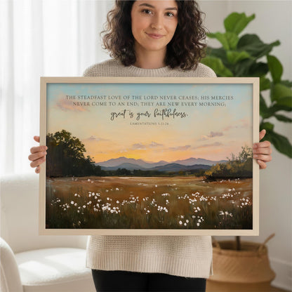 A woman is holding up a framed print of a landscape with mountains and a field of flowers.