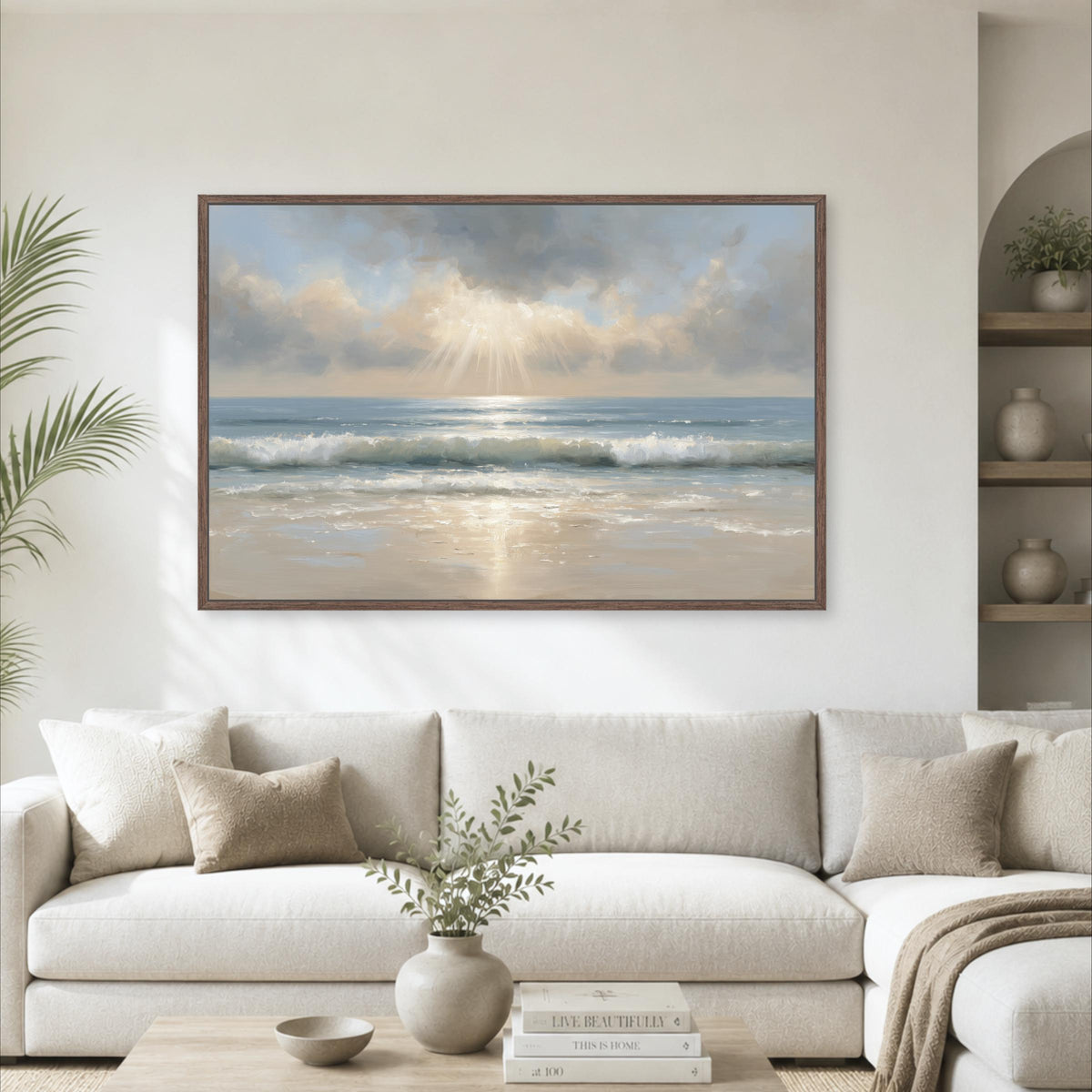 a large framed painting of a serene beach scene with a clear blue sky, ocean waves, and a warm, golden sunset. The painting is hung on a white wall above a comfortable-looking couch with a coffee table in front of it. The room also includes a potted plant, vases, and books, creating a cozy and inviting atmosphere.