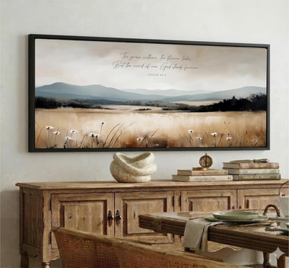 a large framed painting of a serene landscape with mountains, a field of flowers, and a winding path. The painting is hung on a wall above a wooden cabinet, which has a vase and some books on top. The overall scene is peaceful and natural, with the mountains providing a majestic backdrop to the scene.