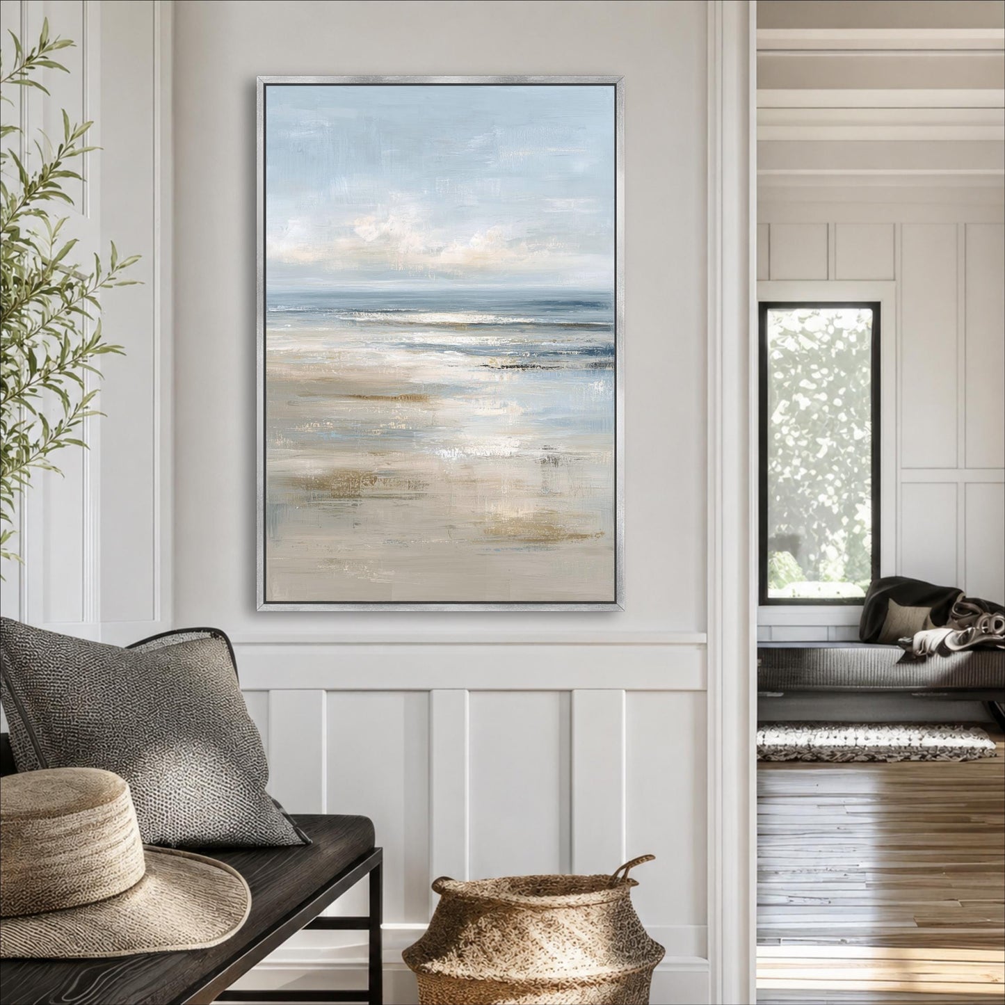 a living room with a large painting of a beach scene hanging on the wall. The painting depicts a serene beach landscape with a calm ocean and a clear sky. The room is decorated with a gray sofa, a wicker basket, and a potted plant, creating a cozy and inviting atmosphere.