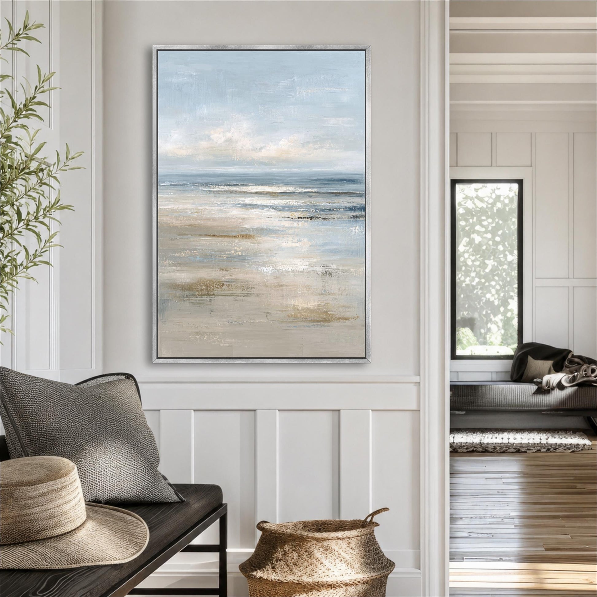 a living room with a large painting of a beach scene hanging on the wall. The painting depicts a serene beach landscape with a calm ocean and a clear sky. The room is decorated with a gray sofa, a wicker basket, and a potted plant, creating a cozy and inviting atmosphere.