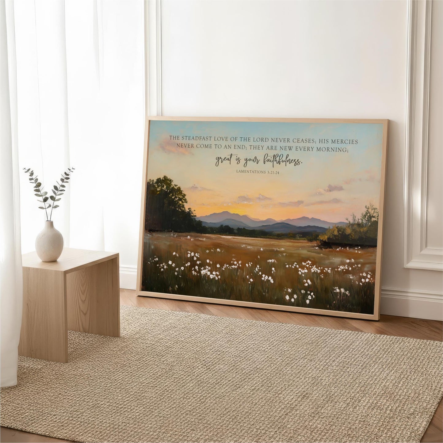 a large framed painting of a serene landscape with a field of flowers and mountains in the background, placed on a wooden floor in a room with a white curtain and a small white vase.