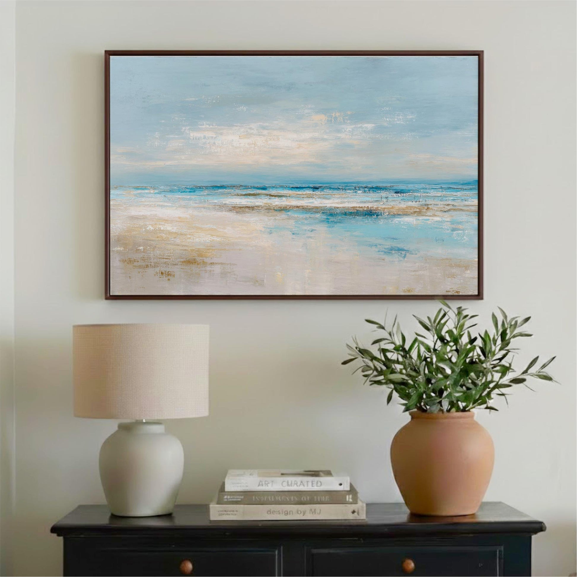 a framed painting of a serene beach scene hanging on a wall above a black dresser with a lamp and a vase of greenery.