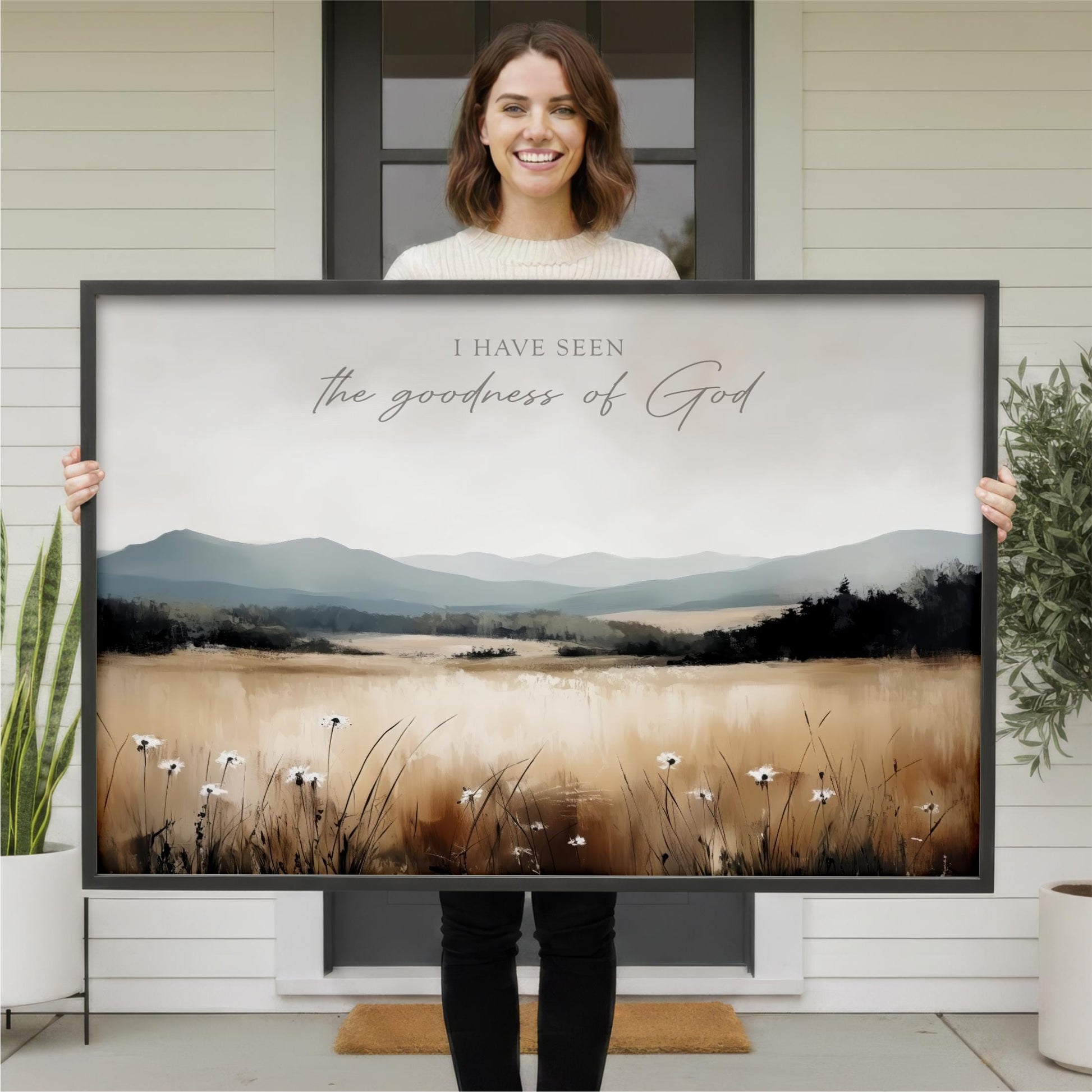 A woman is holding a large framed print of a landscape with mountains and flowers in the foreground.