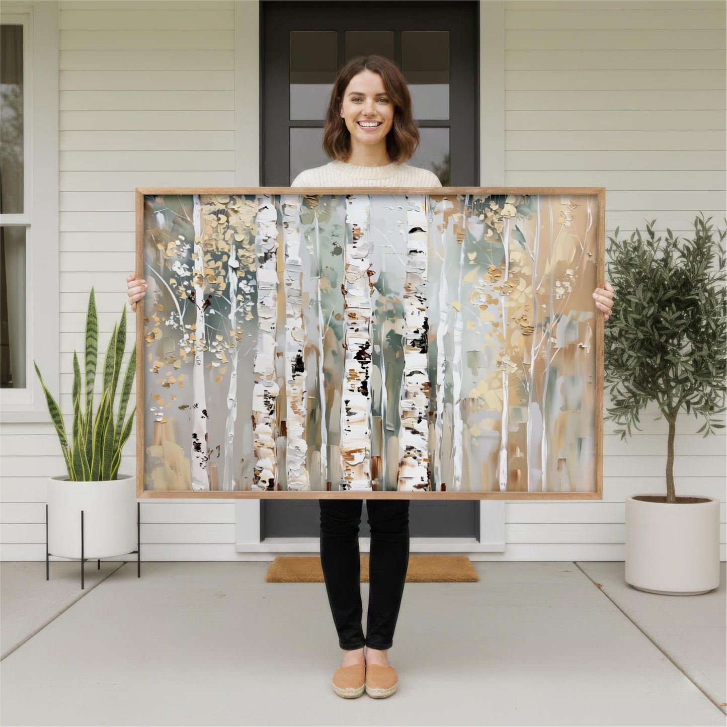 A woman stands in front of a white house, holding a large framed painting of a forest scene.