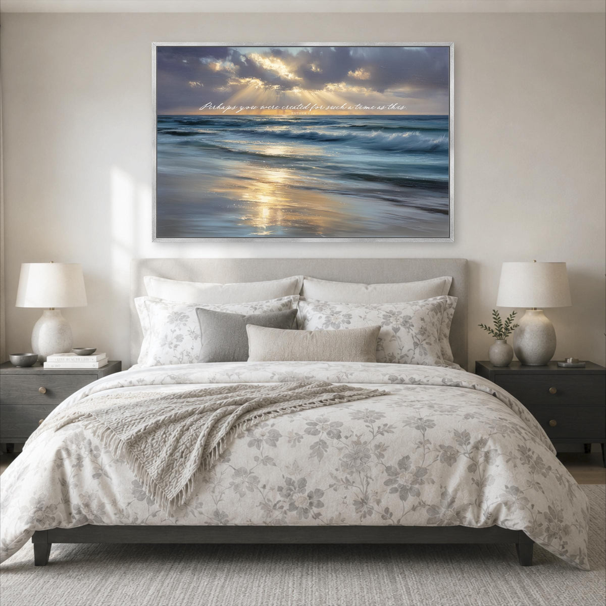 a large, well-made bed with a floral comforter and pillows, flanked by two nightstands with lamps. Above the bed is a large framed picture depicting a serene beach scene with a sunset over the ocean.