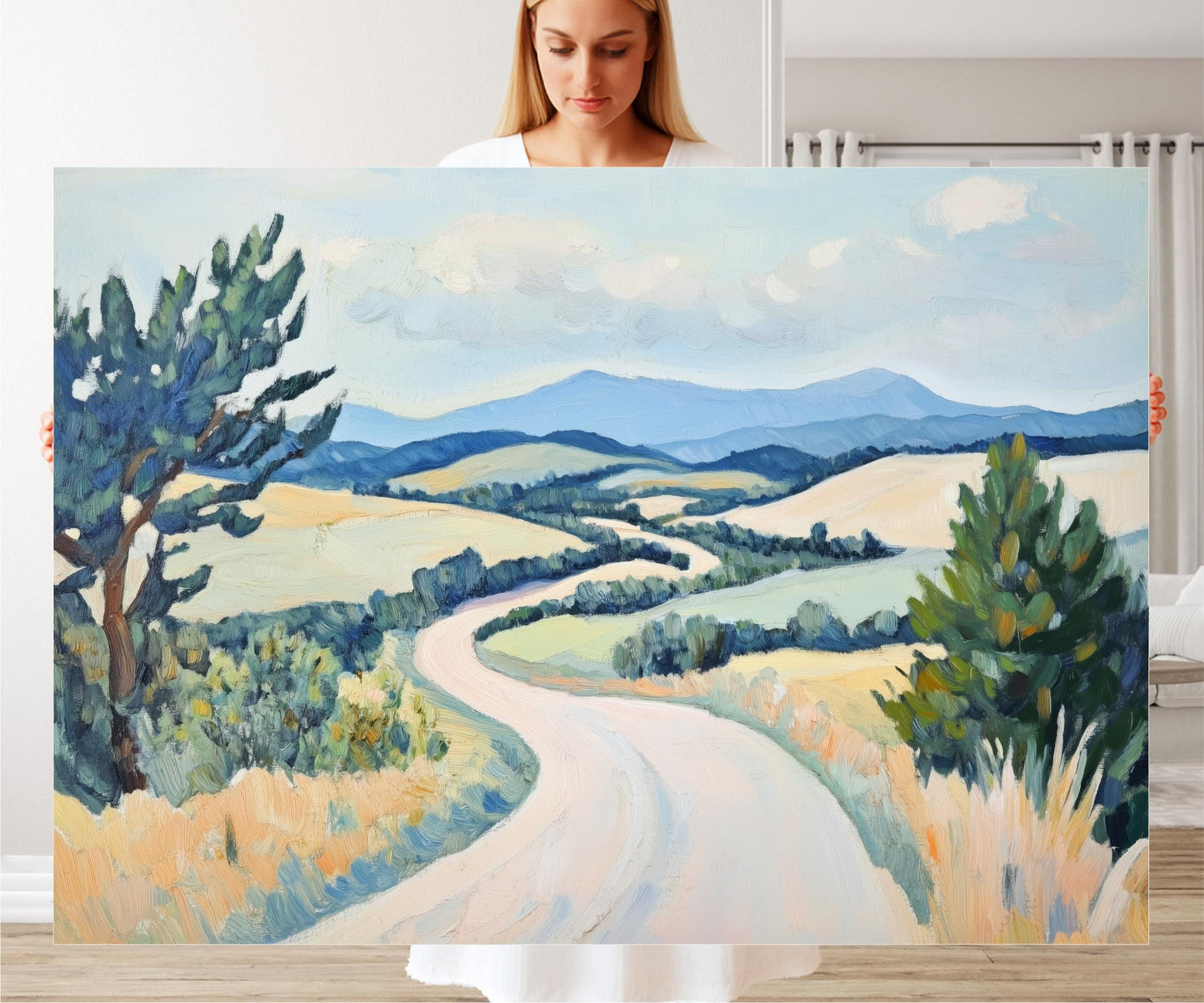 A woman holds a large painting of a winding road through a picturesque countryside landscape, featuring rolling hills, trees, and a blue sky with clouds.